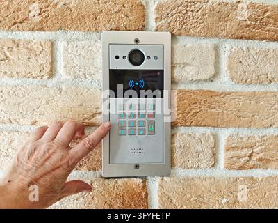 Vue rapprochée d'une main sélectionnant un bouton sur un panneau de communication d'entrée en argent fixé à un mur texturé en pierre, photographié dans un cadre réel. L'ima Banque D'Images