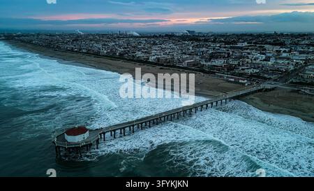 Vue aérienne du Manhattan Beach Pier s'étendant dans l'océan avec des vagues s'écrasant contre les pieux et une silhouette de ville côtière sous un ciel crépusculaire à Ca Banque D'Images