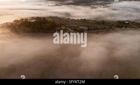 Vue aérienne de la ville historique de Gubbio émergeant d'un épais brouillard matinal avec des collines ondulantes et des cyprès baignés dans la lumière dorée de l'aube à Gubbio Banque D'Images