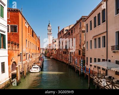 Venise, Italie - 10.07.2016 : cours d'eau pittoresque bordé de façades en terre cuite patinées, de poteaux d'amarrage en bois et d'un haut clocher blanc en arrière-plan Banque D'Images