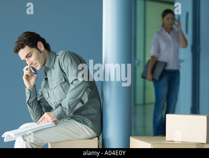 Jeune homme assis sur la boîte en carton à l'aide d'un téléphone cellulaire, jeune femme entrant en arrière-plan Banque D'Images