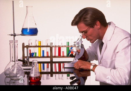 Homme Technicien de laboratoire à la recherche dans un microscope avec tubes à essai et gobelets sur la table à côté de lui Banque D'Images