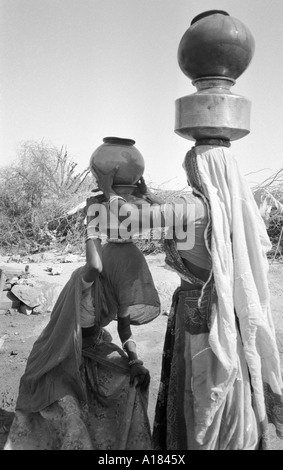 B/W de deux femmes qui chargent des pots d'eau en métal et en argile sur leurs têtes après avoir rempli au puits, une tâche quotidienne pour les femmes rurales au Gujarat, en Inde Banque D'Images