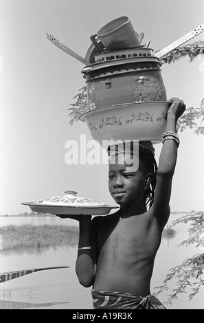 B/W d'une jeune fille de la tribu Bozo équilibrant des casseroles et des poêles de cuisine sur sa tête, Dagua Womina, Mali, Afrique de l'Ouest Banque D'Images