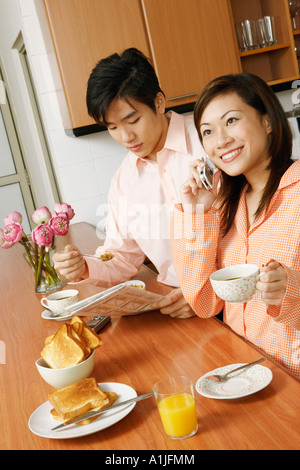 Jeune femme à l'aide d'un téléphone mobile et un jeune homme lisant un journal à un comptoir de cuisine Banque D'Images