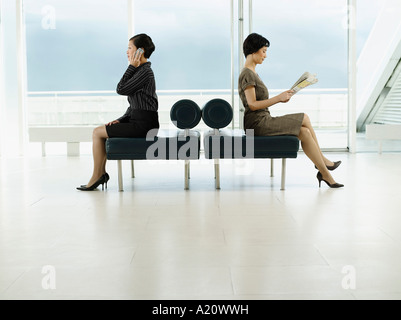 Businesswomen assis sur des bancs, en face de directions opposées, talking on mobile, lisant le journal, profile Banque D'Images