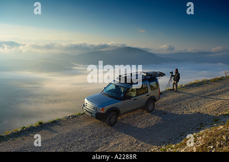 Un photographe travaillant à côté d'un véhicule 4x4 Land Rover au-dessus de la brume couché sur le Piano Grande à l'aube des Monti Sibillini Banque D'Images