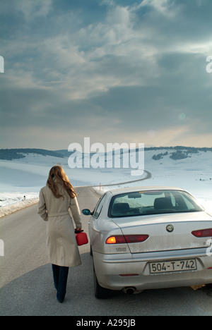 Femme commence la longue marche le long d'une route de campagne isolée en hiver pour trouver du carburant pour sa voiture en panne Banque D'Images