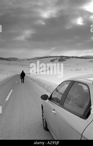 Homme marchant le long d'une route de campagne isolée en hiver pour trouver du carburant pour sa voiture en panne Banque D'Images