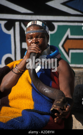 Ndebele woman blowing une corne de kudu en face de doigt painted house, Botshabelo village Ndebele, Afrique du Sud Banque D'Images
