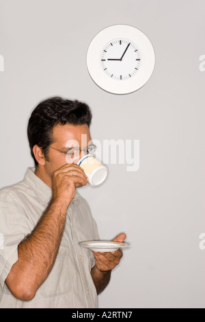 Un jeune homme aux cheveux noirs est debout devant un mur de boire du café ou du thé. L'heure de l'horloge est de cinq (5) minutes après Banque D'Images