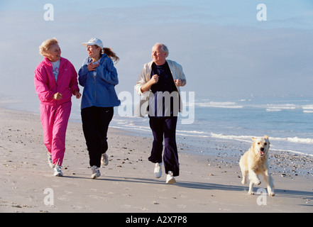 Groupe adultes de trois personnes avec leur chien qui court le long d'une plage. Banque D'Images