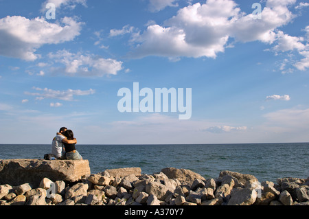 Couple - homme et femme -, assis sur les roches par l'eau, enfermés dans une étreinte. Banque D'Images