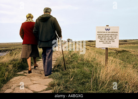 Deux personnes marchant vers la côte avec un panneau d'avertissement de l'érosion côtière Norfolk Broads Angleterre UK Banque D'Images