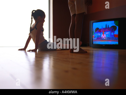 Petite fille assise sur le sol à regarder la télévision, deuxième personne debout à côté d'elle Banque D'Images