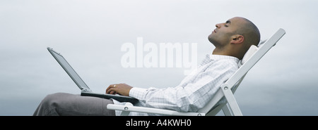 L'homme avec les yeux fermé en chaise longue avec ordinateur portable sur les genoux, ciel en arrière-plan, Vue de côté Banque D'Images