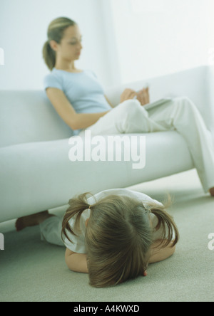 Little girl lying face vers le bas sur le plancher, jeune femme assise sur un canapé, la lecture Banque D'Images