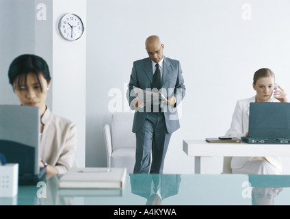 Male executive standing in office reading newspaper, collègues femmes travaillant sur des ordinateurs portables Banque D'Images