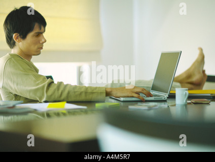 Jeune homme assis à table avec pieds, using laptop Banque D'Images