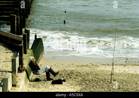 Un homme âgé assis sur la plage, dormir, avec un chien à côté de lui et la canne à pêche courte distance, Suffolk, UK Banque D'Images