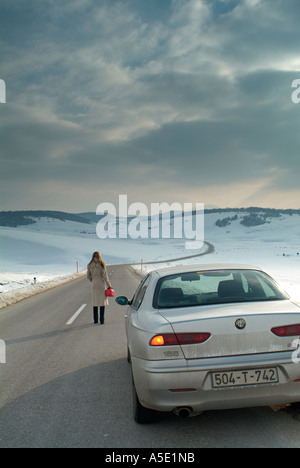 Femme marche le long d'une route de campagne isolée en hiver pour trouver du carburant pour sa voiture en panne Banque D'Images