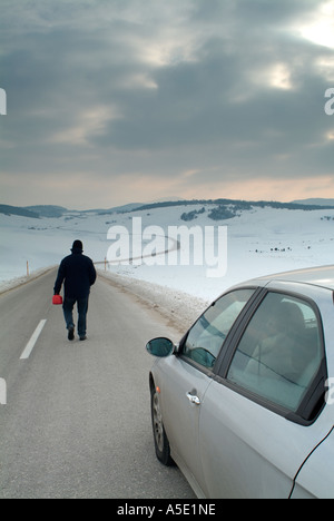 Homme quitte son conjoint dans la voiture pour marcher le long d'une route isolée dans une vallée couverte de neige à la recherche de carburant Banque D'Images