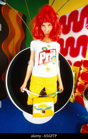 Femme en fauteuil portant perruque rouge, pantalon jaune et Pril t-shirt des années 70, sac à main, portefeuille avec décoration de la 70i Banque D'Images