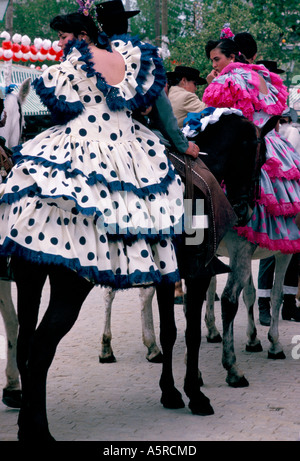 Les JEUNES FEMMES PORTANT DES ROBES DE FLAMENCO SEVILLANAS CIRCONSCRIPTION DEUX JUSQU'À L'ARRIÈRE DES CHEVAUX montés par des hommes en tenue traditionnelle WIT Banque D'Images