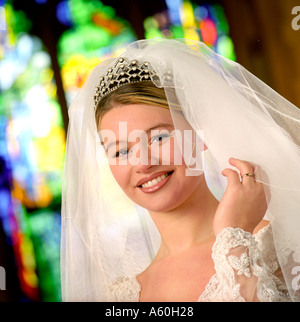 Mariée en robe nuptiale traditionnelle et voile wearing wedding ring sourit à l'appareil photo à l'intérieur de l'église vitrail derrière Banque D'Images