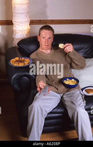 Homme assis sur setee entouré de junk food holding television remote controller Banque D'Images