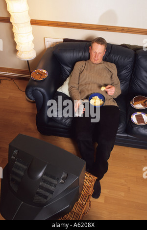 Homme assis sur setee entouré par la malbouffe devant la télévision holding remote controller Banque D'Images