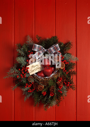 Couronne de fruits rouges et de conifères avec des pommes rouges et ruban tartan sur porte rouge Banque D'Images