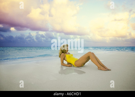 Jolie jeune femme blonde se prélasser sur la plage en maillot jaune aux Maldives dans l'Océan Indien au coucher du soleil Banque D'Images