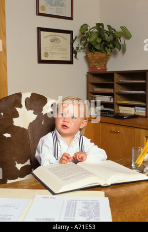 Jeune garçon portant des bretelles est assis dans sa chaise de bureau père juridique avec les livres et les papiers sur le bureau en face de lui Banque D'Images