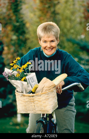 La haute dame assise sur un vélo avec un panier de fleurs contenant une bouteille de vin et un morceau de pain à l'intérieur de son panier Banque D'Images