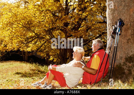 Senior couple sitting in meadow, man leaning on tree, side view Banque D'Images