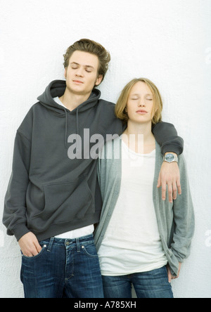 Jeune couple leaning against wall, bras de l'homme autour de l'épaule de la femme, aussi bien fermer les yeux Banque D'Images