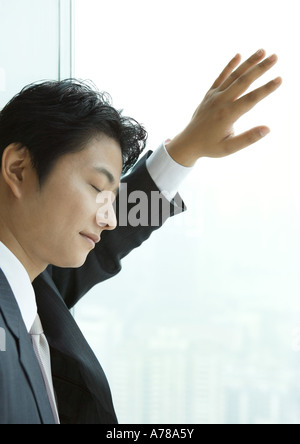 Businessman leaning against window de grande hauteur, les yeux fermés Banque D'Images