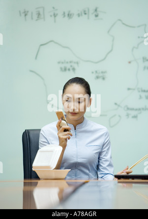 Businesswoman sitting at desk de manger les plats à emporter et using cell phone Banque D'Images