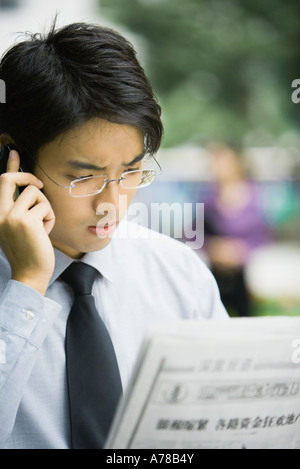 Businessman reading newspaper et using cell phone, fronçant Banque D'Images