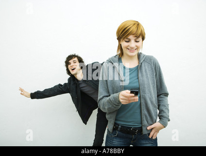 Young woman holding cell phone alors que jeune homme derrière elle des danses, fond blanc Banque D'Images