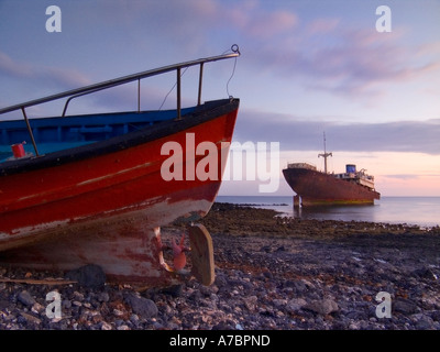 Naufrage de rouille se trouve en eau peu profonde dans la crique rocheuse avec bateau de pêche rouge abandonnés en premier plan sur la plage de galets Banque D'Images