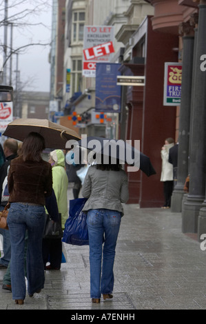 Deux jeunes femmes portant vertical parapluies humides arpentant une rue commerçante dans le centre-ville de Belfast à pied à partir de l'appareil photo Banque D'Images