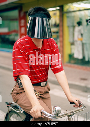 Un chinois de Shanghai homme porte un bouclier de protection d'empêcher le soleil de son visage alors qu'il chevauche son vélo dans la rue Banque D'Images