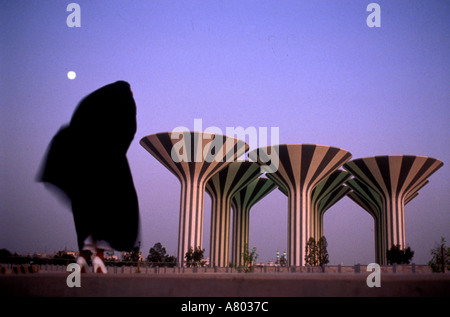 Arab woman in black burka blowing in wind révèle des chaussures blanc près de Koweït City Tours à l'eau comme à la tombée de la lune se lève Banque D'Images