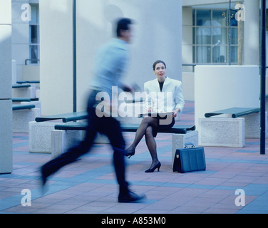 L'homme qui traverse city mall passé femme assise sur un banc à l'extérieur de bâtiment de bureaux. Banque D'Images