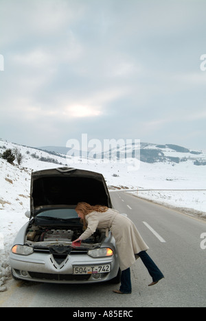 Jeune femme à la recherche sous le capot de sa voiture après avoir été coincé sur une route isolée en hiver après une rupture Banque D'Images