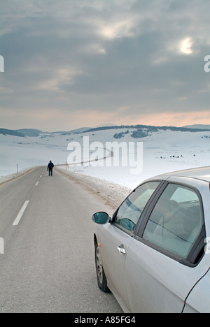 Homme quitte son conjoint dans la voiture pour marcher le long d'une route isolée dans une vallée couverte de neige à la recherche de carburant Banque D'Images