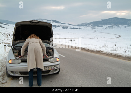 Jeune femme à la recherche sous le capot de sa voiture après avoir été coincé sur une route isolée en hiver après une rupture Banque D'Images