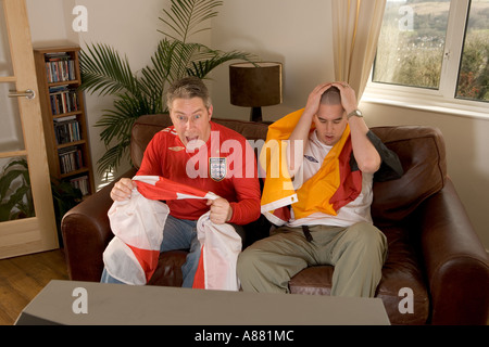 Parution du modèle. L'Angleterre et l'Allemagne de football fans assis sur le canapé à regarder la télévision que l'Angleterre gagne et perd de l'Allemagne. Banque D'Images